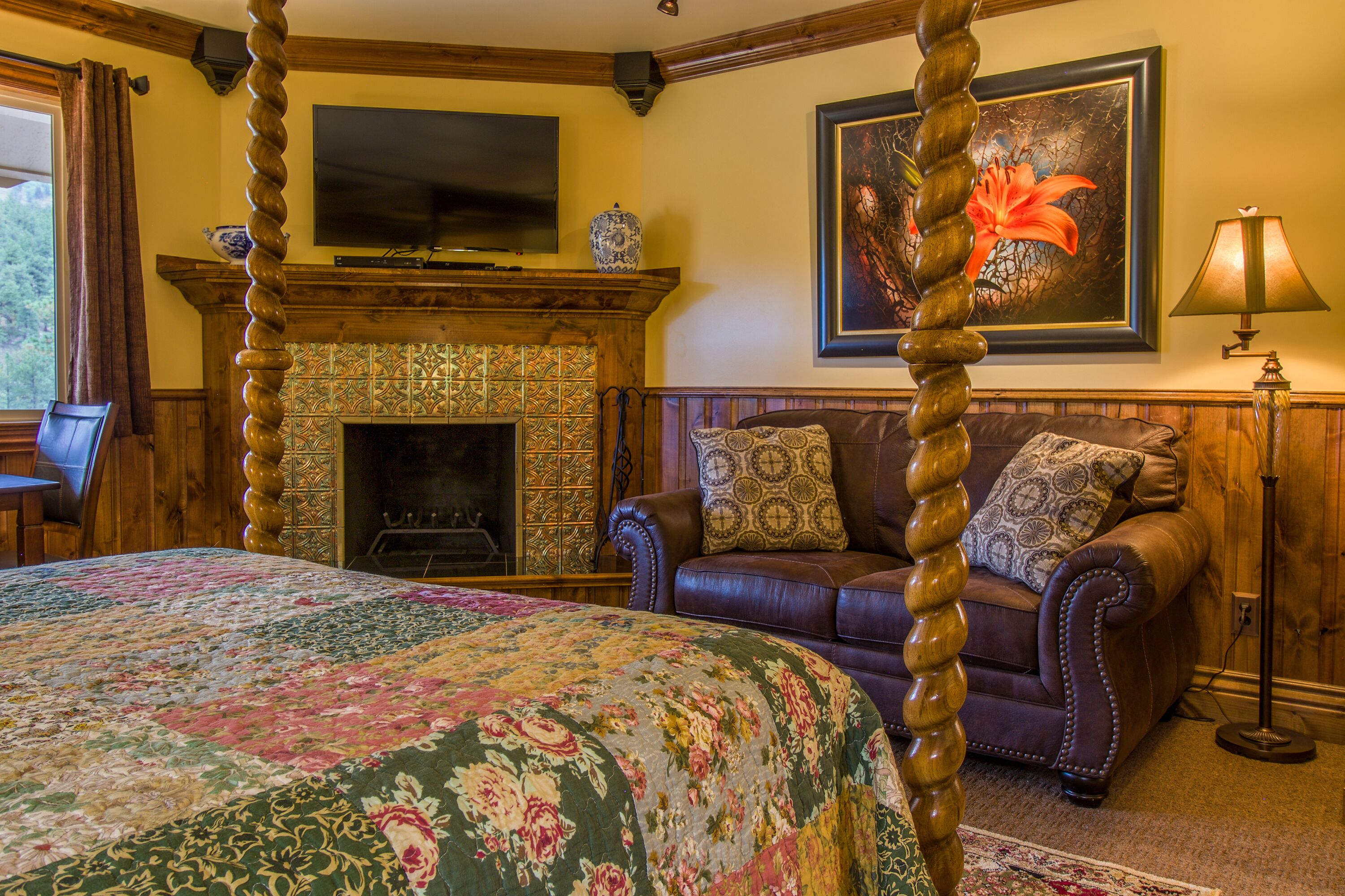 A bedroom with a four-poster bed, a fireplace, a leather sofa, and a television on the wall.