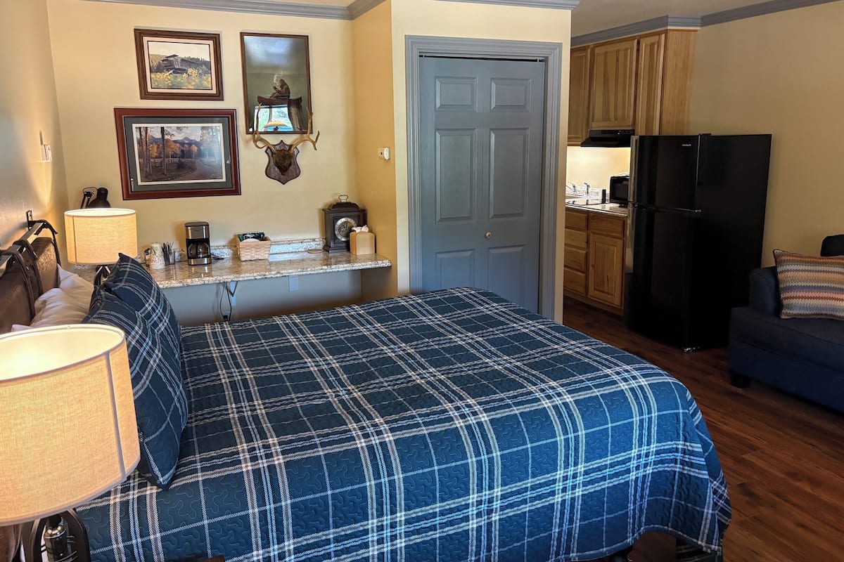 Un dormitorio con cama, mesita de noche, lámpara, cómoda, espejo y puerta que da a una cocina.
