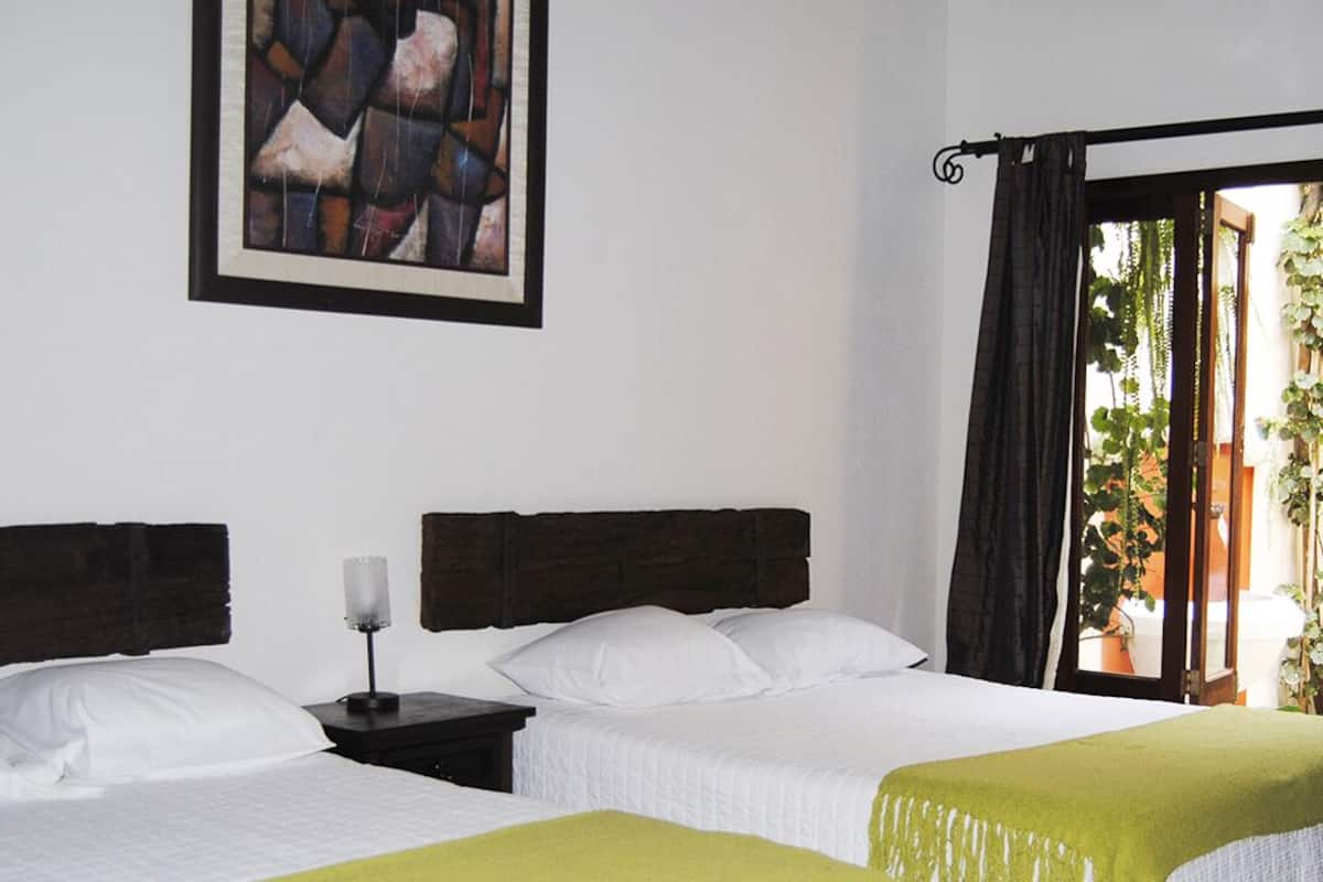 A hotel room with two beds, a nightstand with a candle, a painting on the wall, and a view of greenery outside.