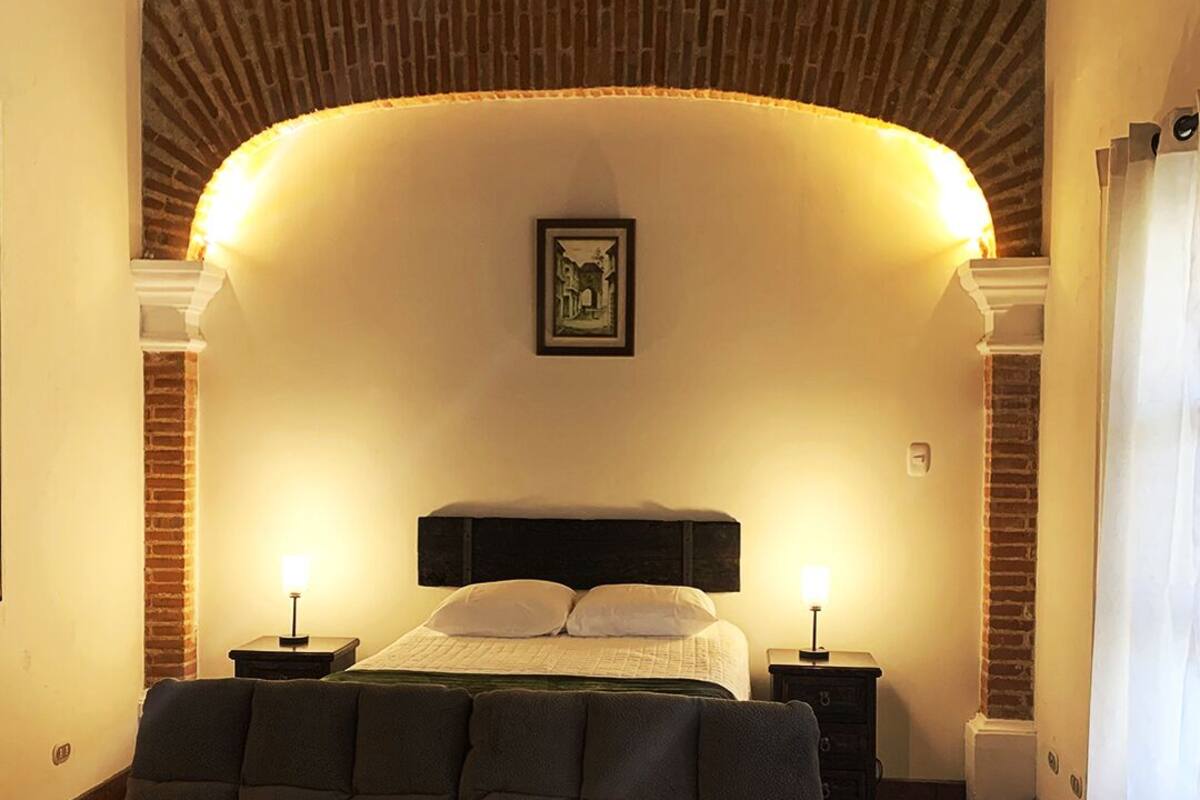 A bedroom with a brick wall, a bed with white linens, two bedside tables with lamps, and a large cushioned headboard.