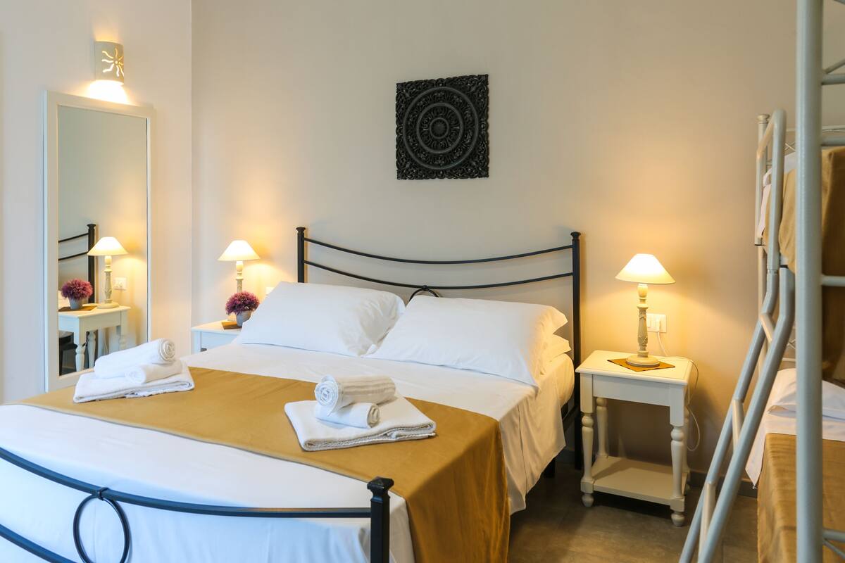 A neatly made bed with white linens and a brown bedspread, two bedside tables with lamps, a wall-mounted decorative piece, and a bunk bed structure to the right.