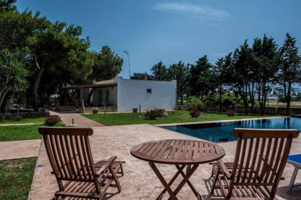 Zona de piscina amb un edifici blanc, dues cadires de fusta i una taula.