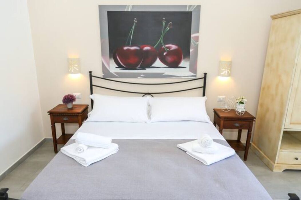 A neatly made bed with white linens, two bedside tables with lamps, and a painting of cherries on the wall.