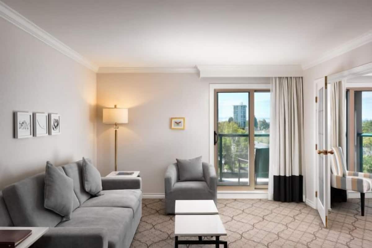 A hotel room with a grey sofa, a smaller armchair, a white coffee table, a floor lamp, and a view of the city through large windows.