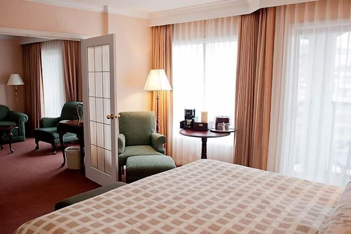 A hotel room with a large bed, a green armchair, a small table with a lamp, and a view of the outdoors through large windows.