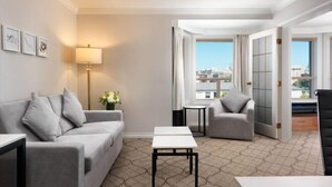 Ein Hotelzimmer mit Sofa, Sessel, kleinen Tischen, einer Lampe und Blick auf Gebäude durch das Fenster.