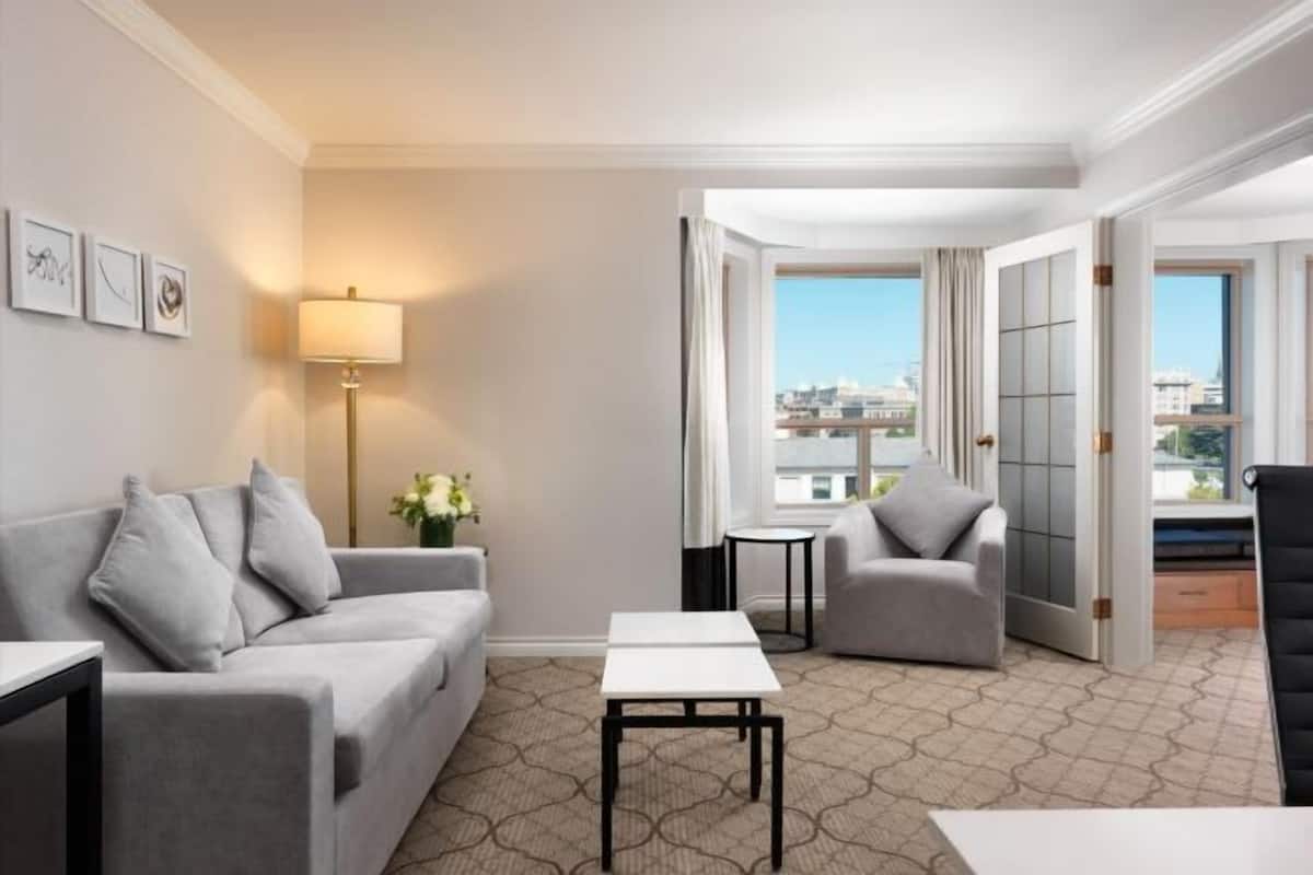 A hotel room with a sofa, armchair, small tables, a lamp, and a view of buildings through the window.