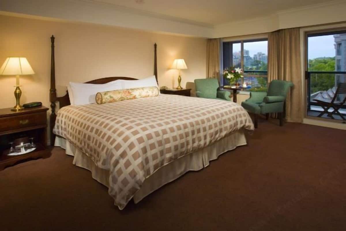 A hotel room with a large bed, two bedside tables with lamps, a green armchair, a small table with a vase, and a balcony with a view.