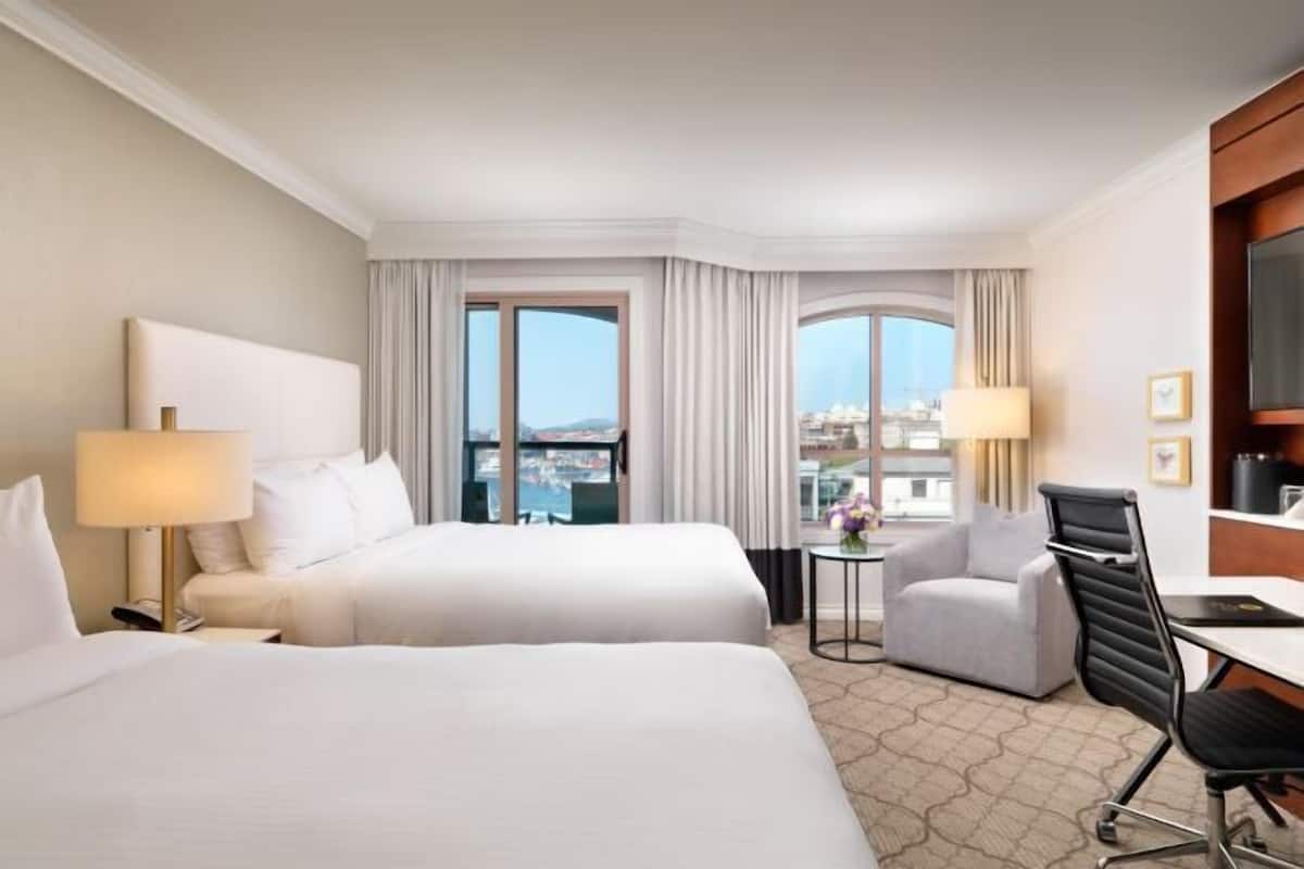 A hotel room with two beds, a desk, a chair, a TV, and a view of the city.