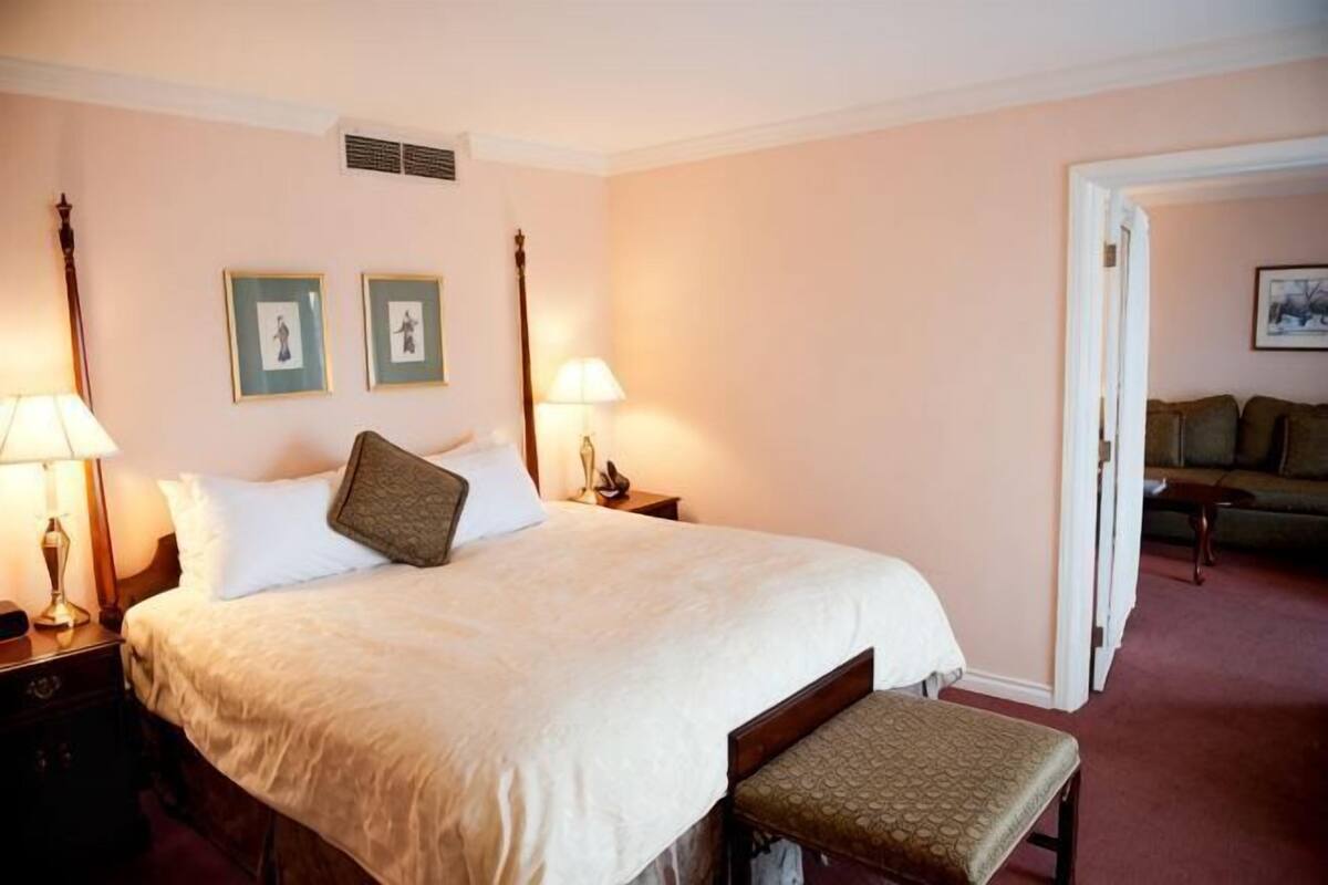 A hotel room with a large bed, bedside tables, a bench, and a door leading to another room.