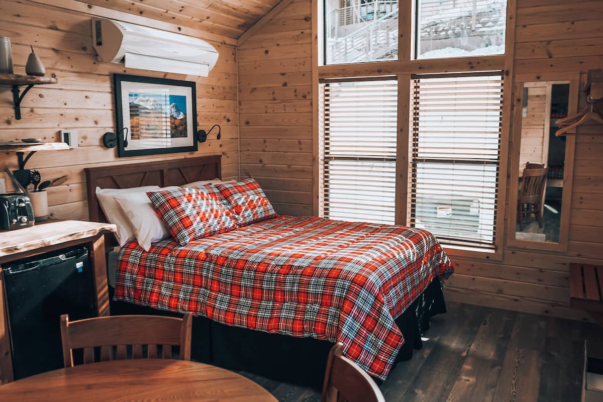 A cozy cabin-style room with a bed, a small table, and a chair. There is a window with blinds, a framed picture on the wall, and a wooden ceiling.