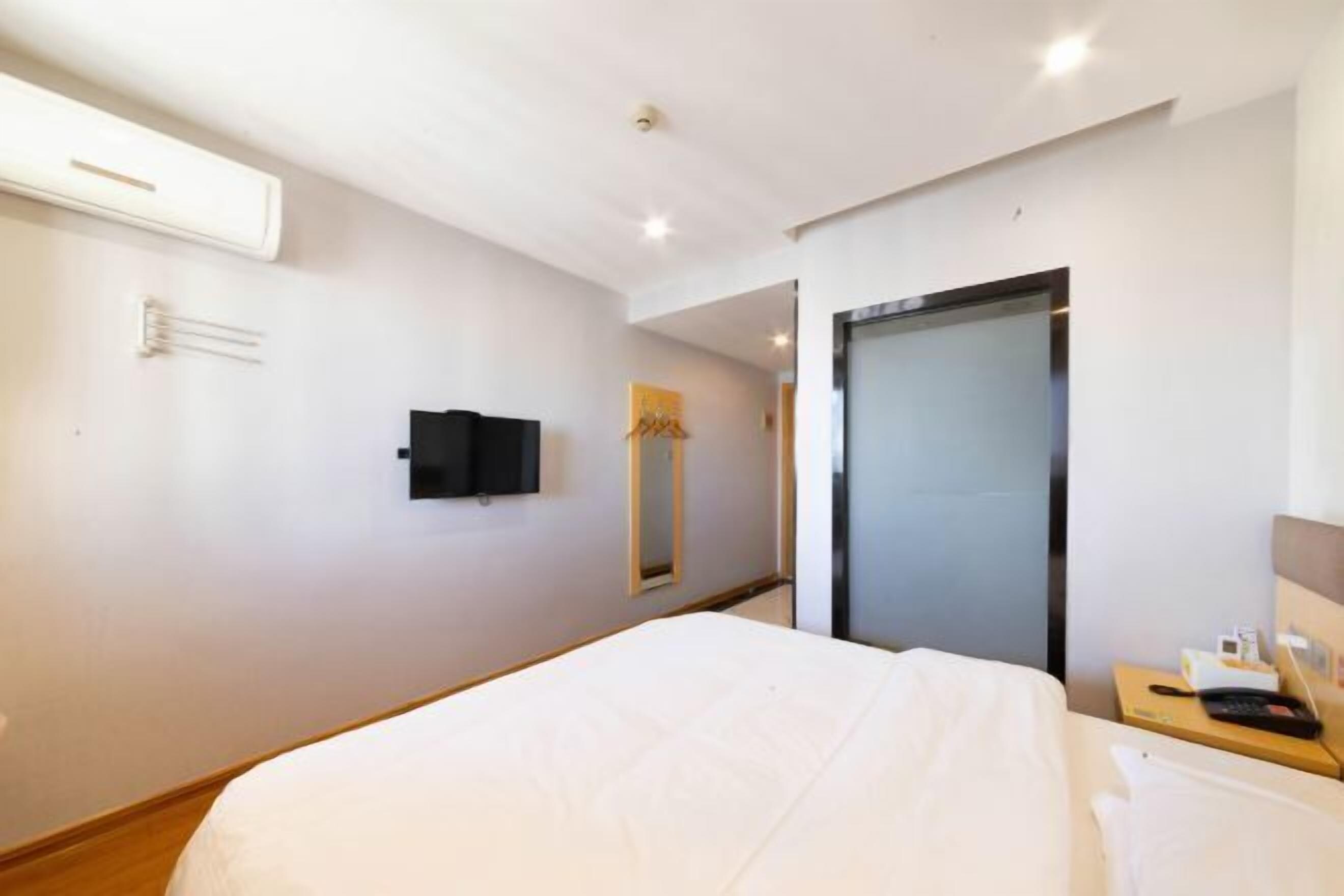 A hotel room with a bed, a bedside table with a phone and a lamp, a wall-mounted TV, and a door with a frosted glass panel.
