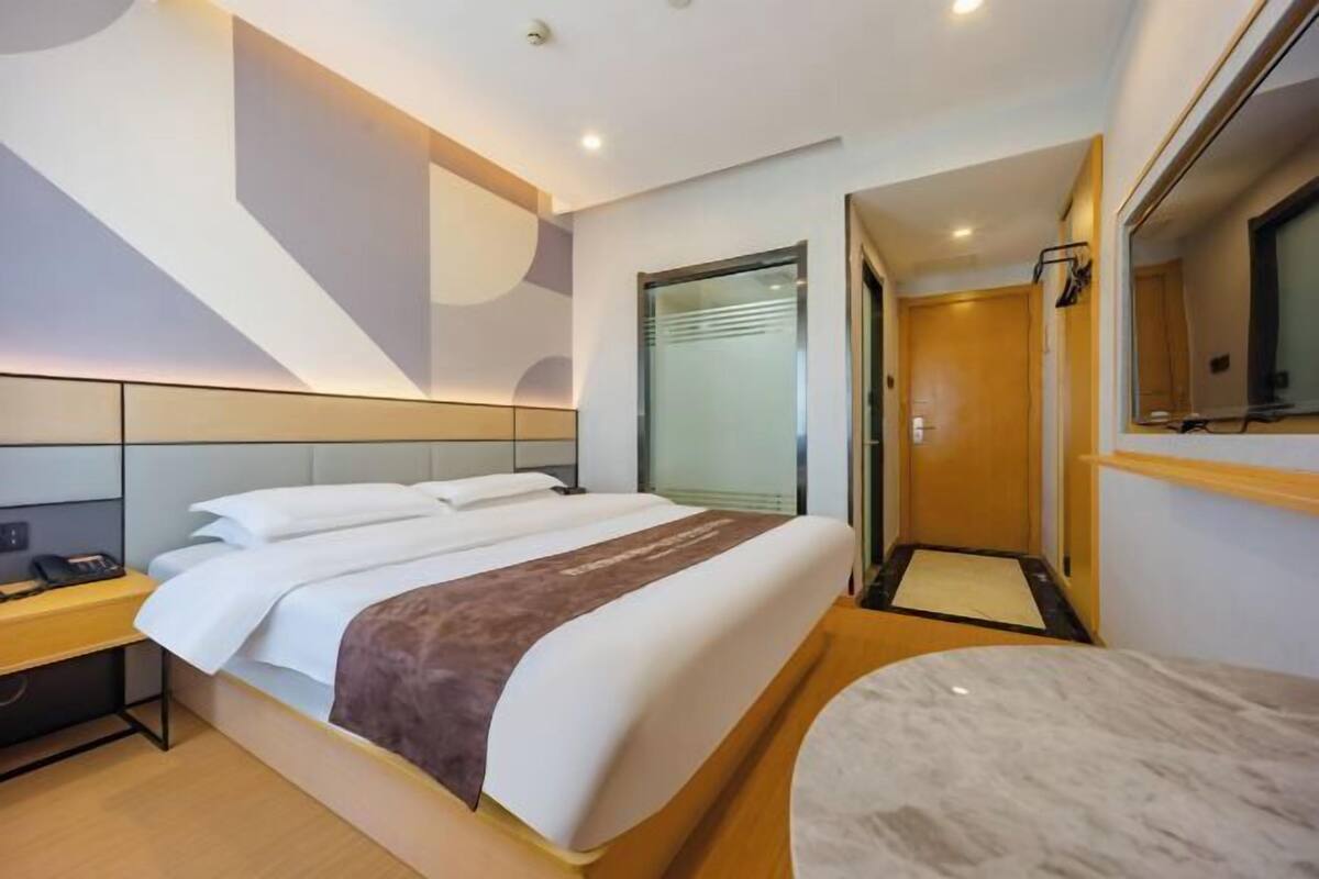 A modern hotel room with a large bed, a bedside table, a mirror, and a door leading to a bathroom.