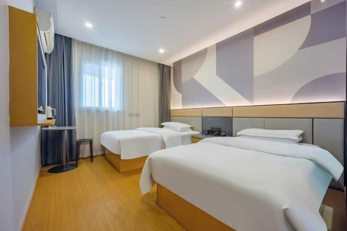 A hotel room with two beds, a small table, a chair, and a large window with curtains.