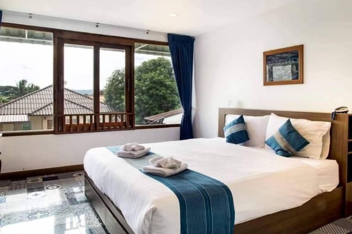 A hotel room with a large bed, a view through a window, and a painting on the wall.