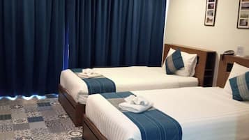 A hotel room with two beds, a large window with dark curtains, a patterned carpet, and a small bedside table with a lamp.