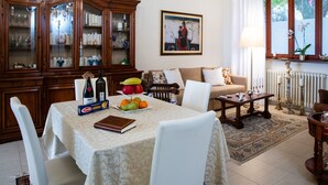 A dining area with a table set for two, a cabinet with glass doors, a sofa, and a window with curtains.
