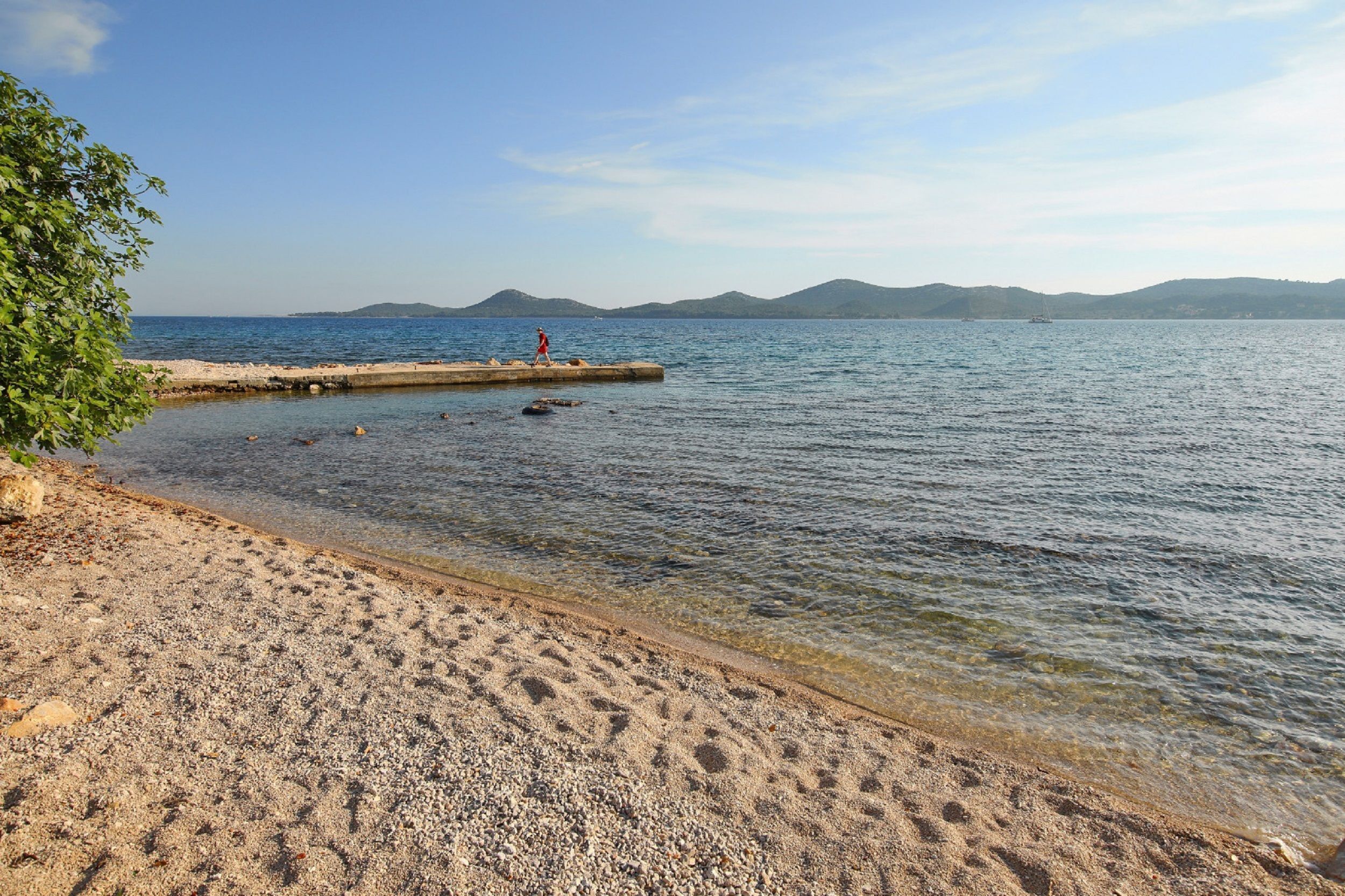 A beach with clear water, a person walking on a pier, and distant mountains.