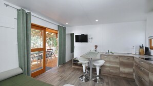 A modern kitchen with a wooden countertop, two white bar stools, a TV mounted on the wall, and a sliding glass door leading to an outdoor area with a wooden deck and chairs. - Mobile Home Smaragd (Biograd)
