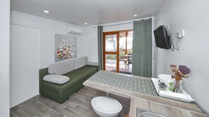 A compact living space with a green sofa, a small table, a white toilet, and a TV mounted on the wall. - Mobile Home Smaragd (Biograd)