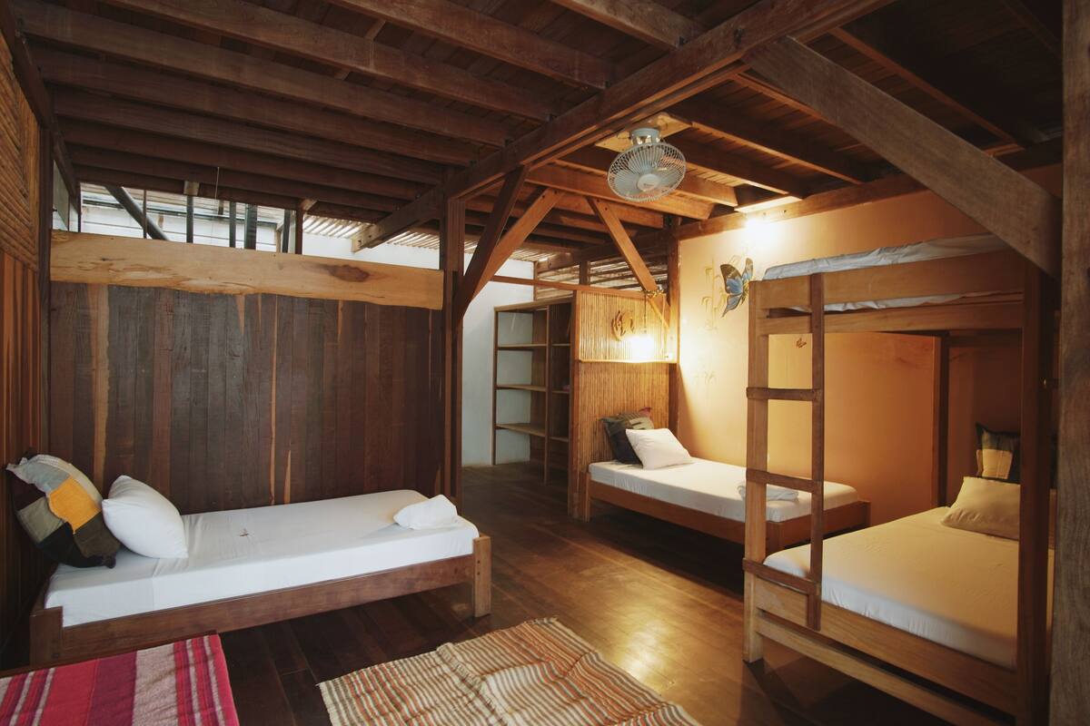A rustic wooden room with two bunk beds and a single bed, wooden beams on the ceiling, and a ladder attached to one of the beds.