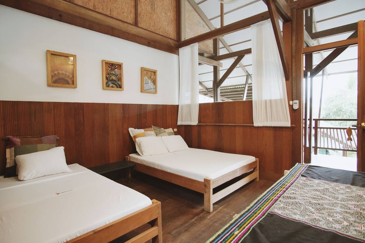 A room with two wooden beds, white bedding, wooden panel walls, and a view through a large window.