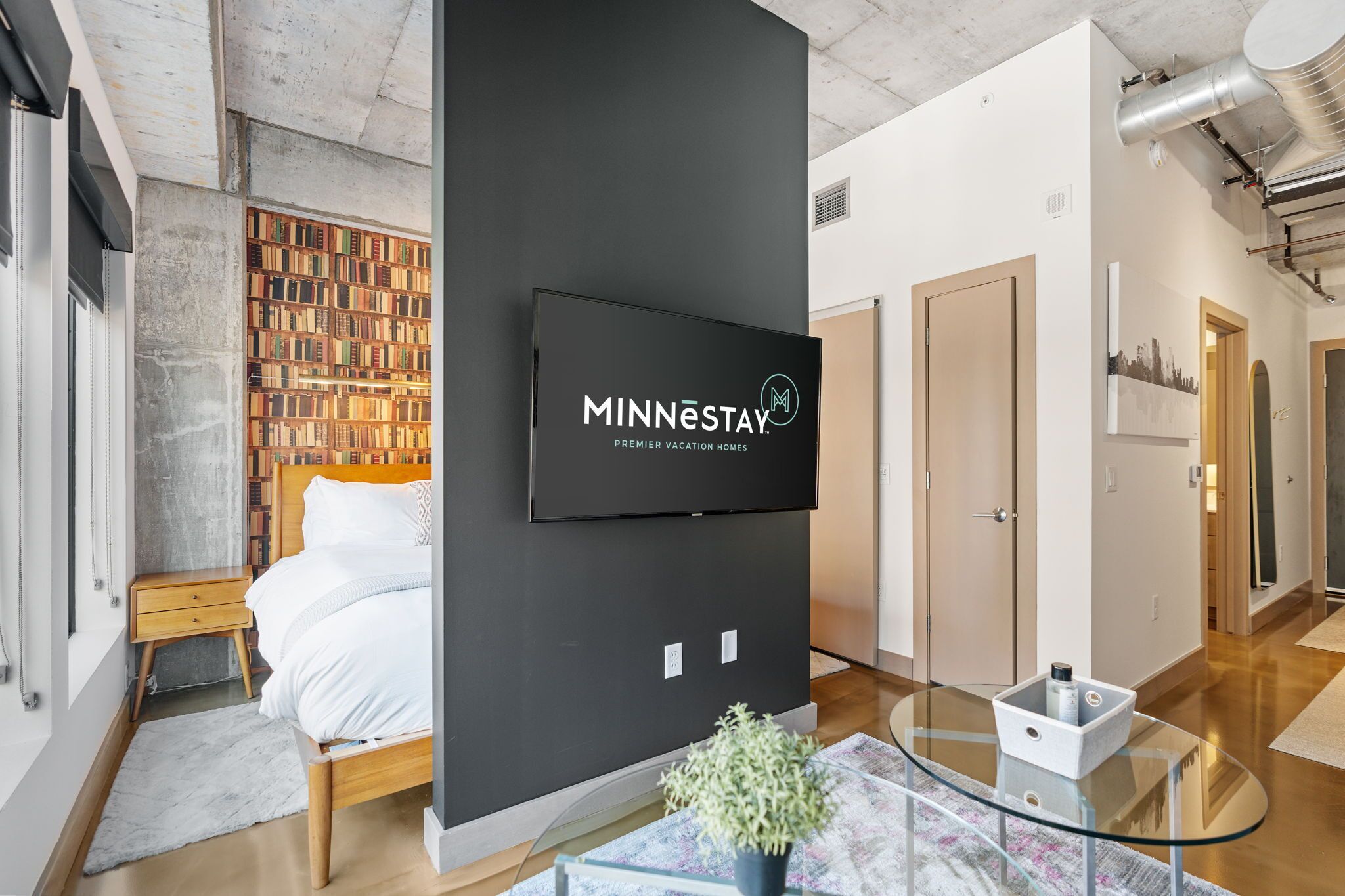 Una habitación de hotel moderna con una gran televisión de pantalla plana montada en una pared negra, una cama de madera, una mesa de centro de cristal y una estantería llena de libros.
