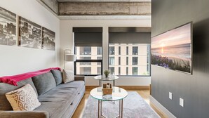 Een moderne woonkamer met een grijze bank, een glazen salontafel en grote ramen met uitzicht op gebouwen en een zonsondergang.
