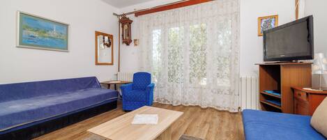 A living room with a blue sofa, a wooden coffee table, a blue armchair, a television on a wooden stand, and a window with floral curtains.