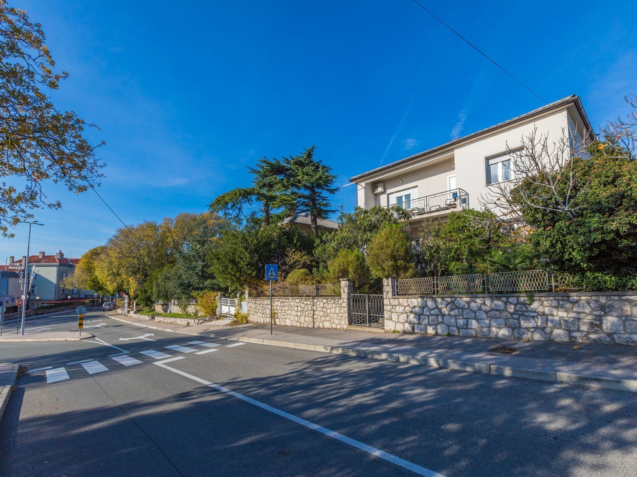 A residential street with a two-story house, a stone wall, and a pedestrian crossing.