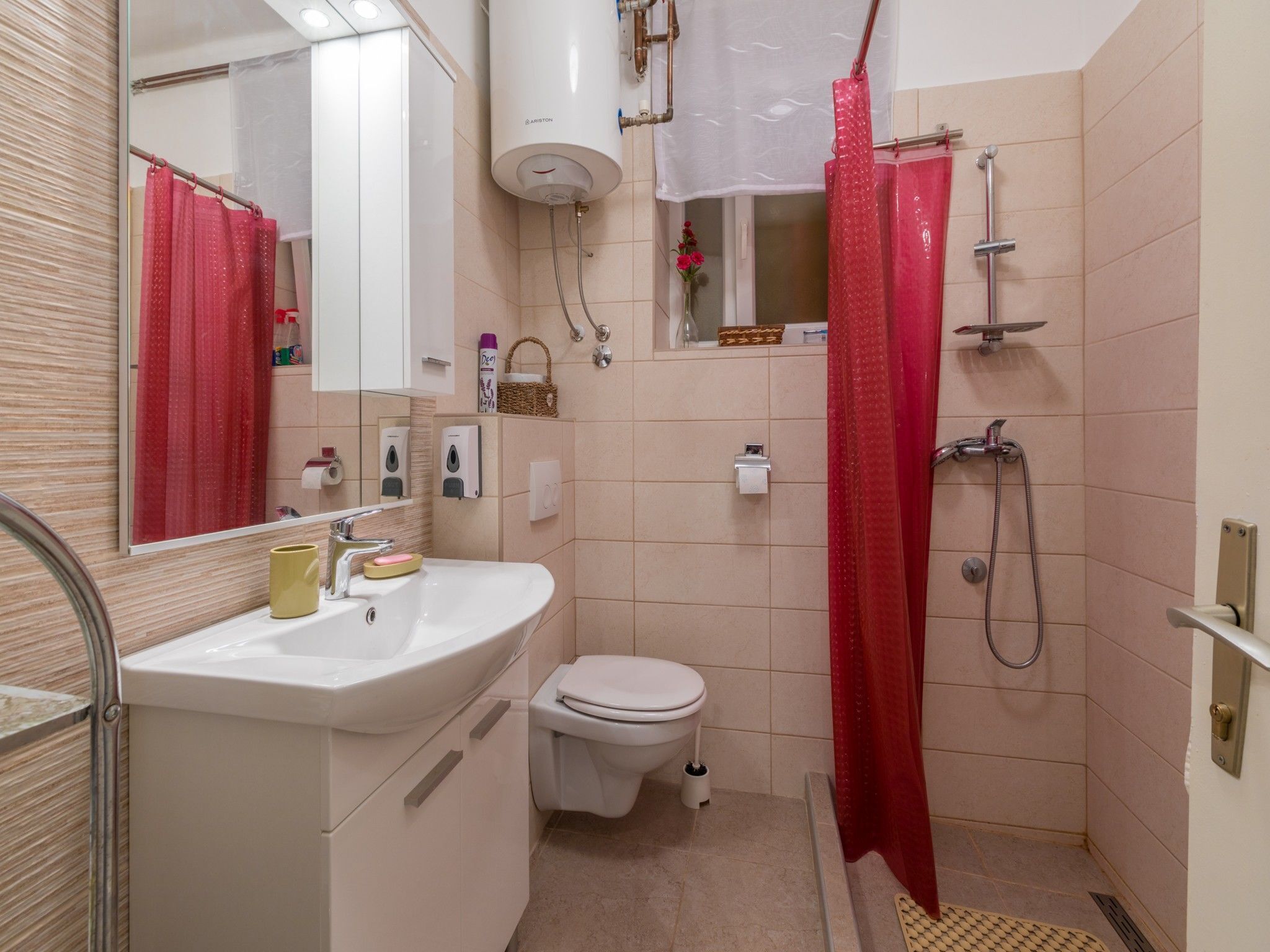 Un baño con un lavamanos blanco, un inodoro, una ducha con cortina roja y un calentador blanco en la pared.