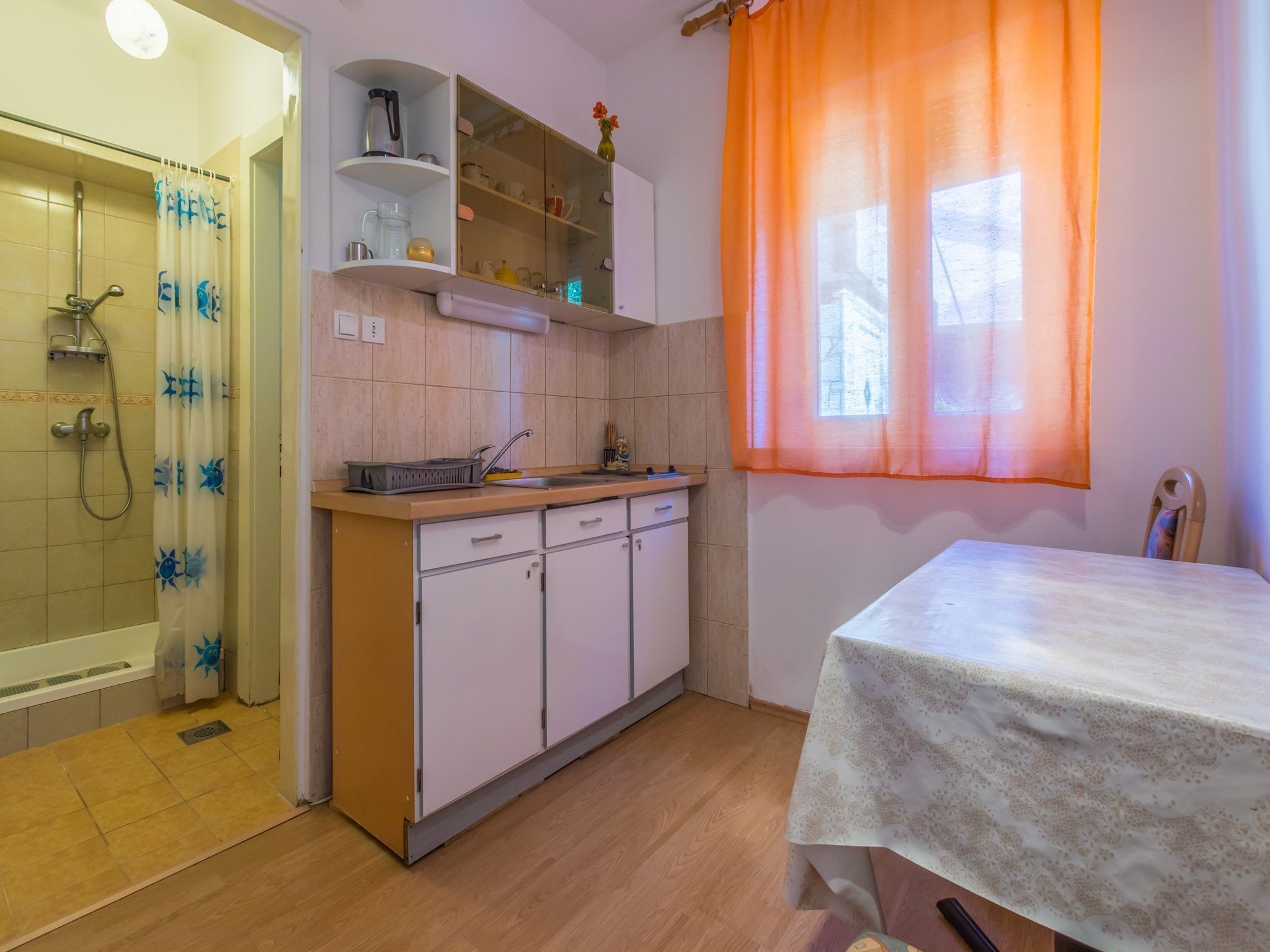 Une cuisine de taille réduite, équipée d’un évier, de placards et d’une fenêtre agrémentée de rideaux orange.