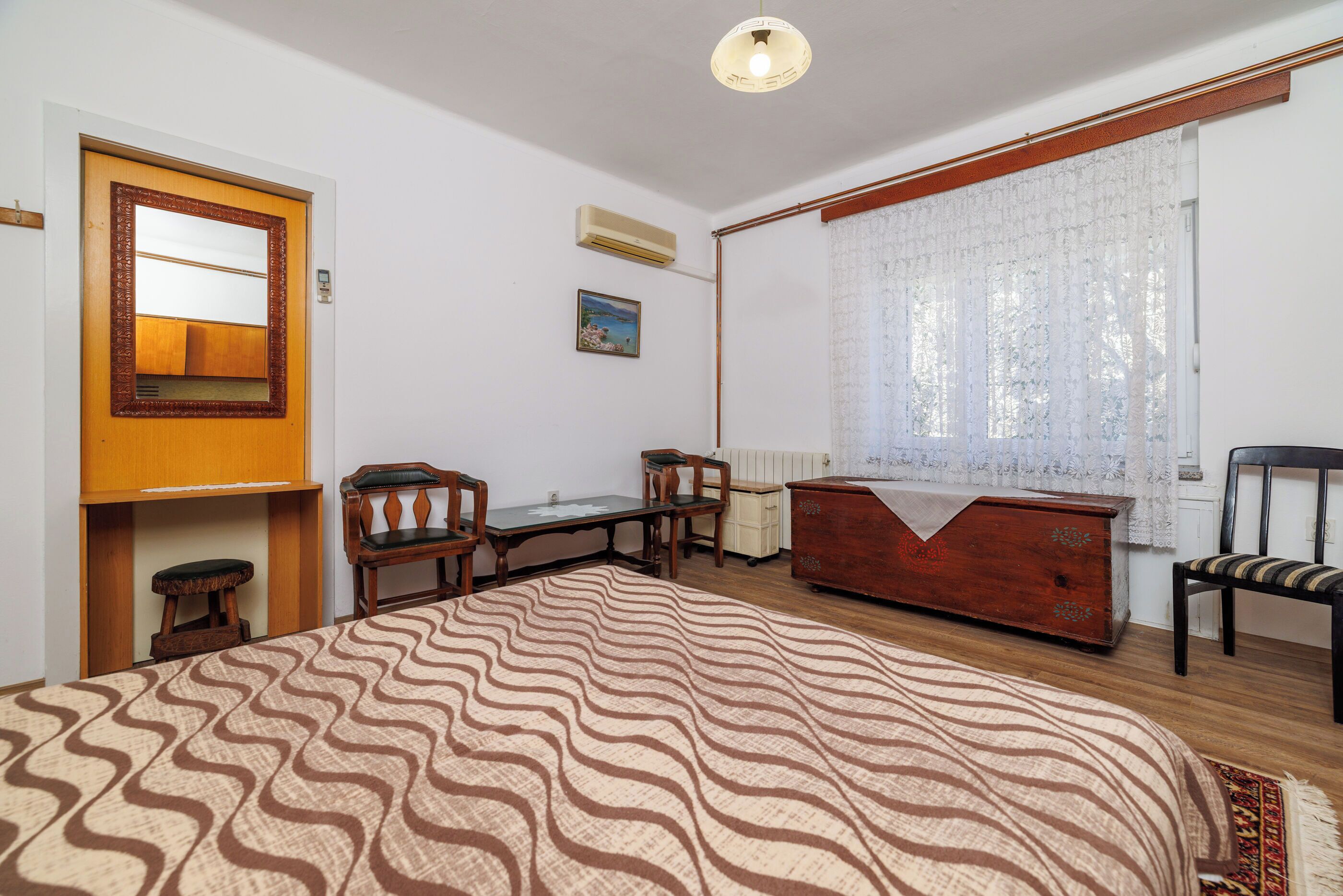 A bedroom with a bed, a wooden dresser, a desk, two chairs, a window with curtains, and a wall-mounted air conditioner.
