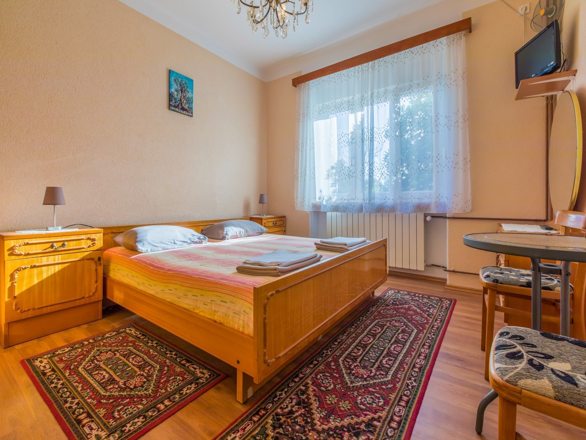 A bedroom with a wooden bed, two bedside tables with lamps, a desk with a chair, a large window with sheer curtains, and a chandelier.