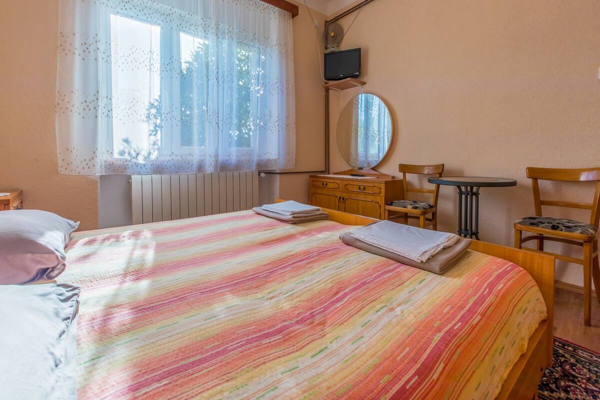 Een slaapkamer met een bed, een dressoir, een stoel, een klein tafeltje, een spiegel en een raam met dunne gordijnen.