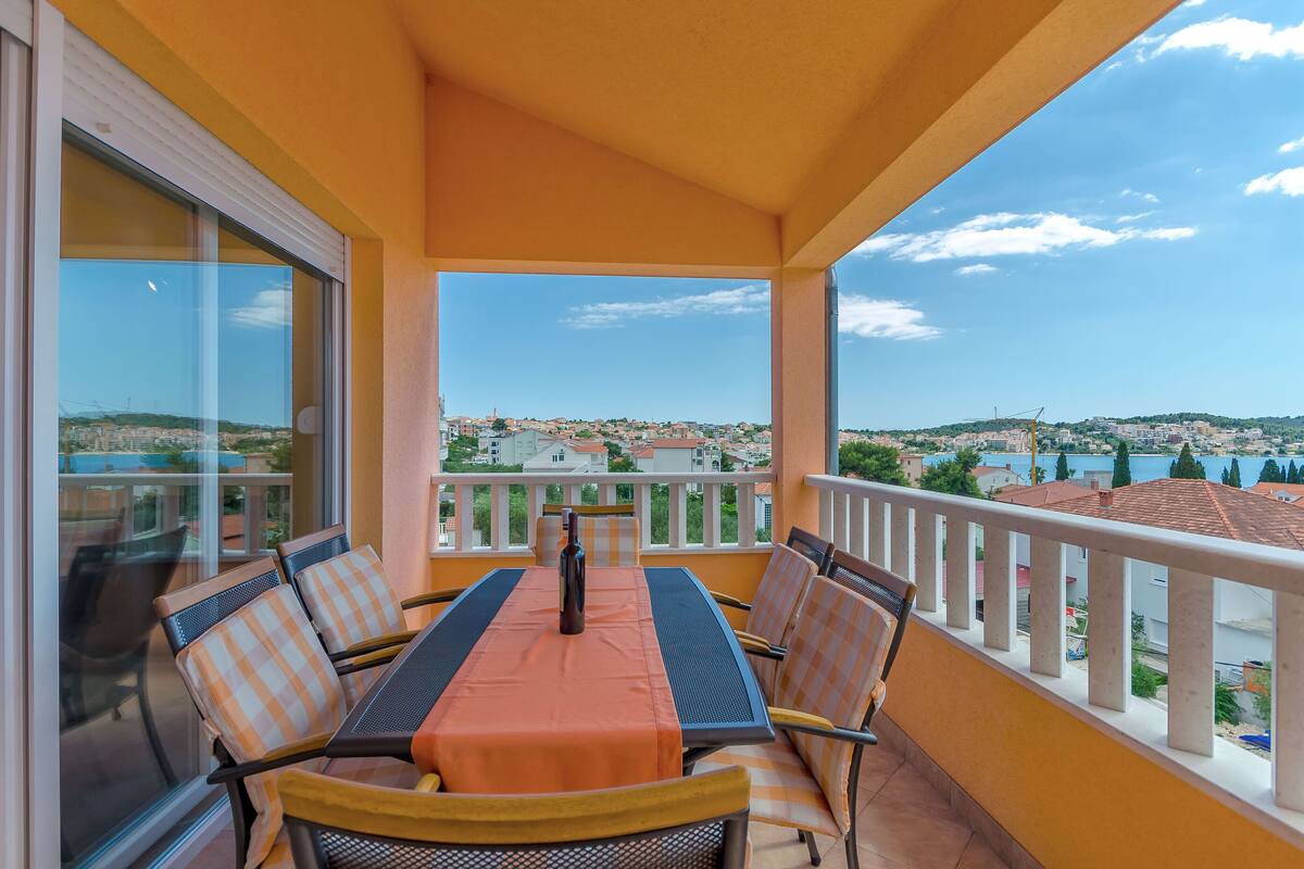 A balcony with a table and chairs, overlooking a coastal town.