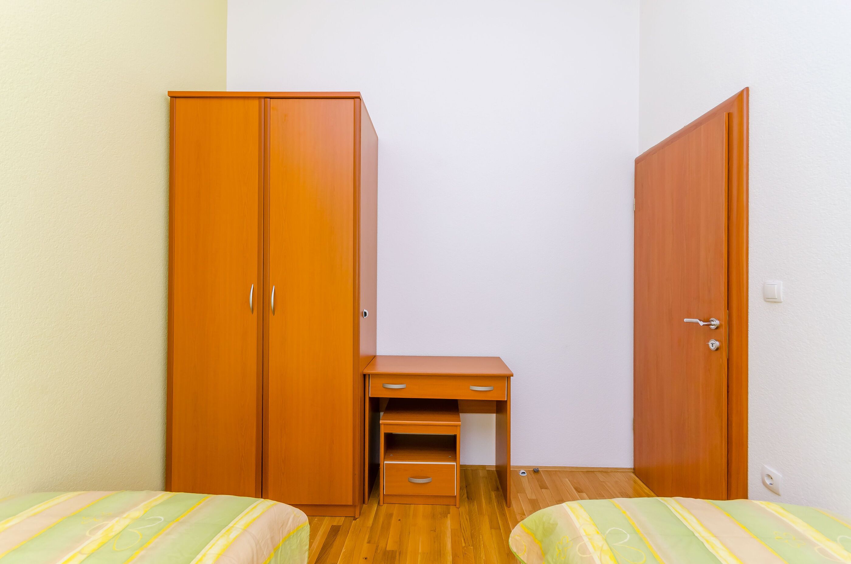 A small, neatly arranged bedroom with a wooden wardrobe, a desk with drawers, and two beds with striped bedding.
