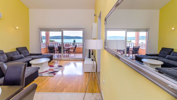 A modern living room with a large sectional sofa, a coffee table, and a dining area with chairs. A large mirror reflects the room and a balcony with a view of the sea. - Apartments Melita (Okrug Gornji)