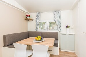 A dining area with a wooden table, white chairs, a bench, and a window with curtains. - Mobile Home Pecten 02 (Pakostane)