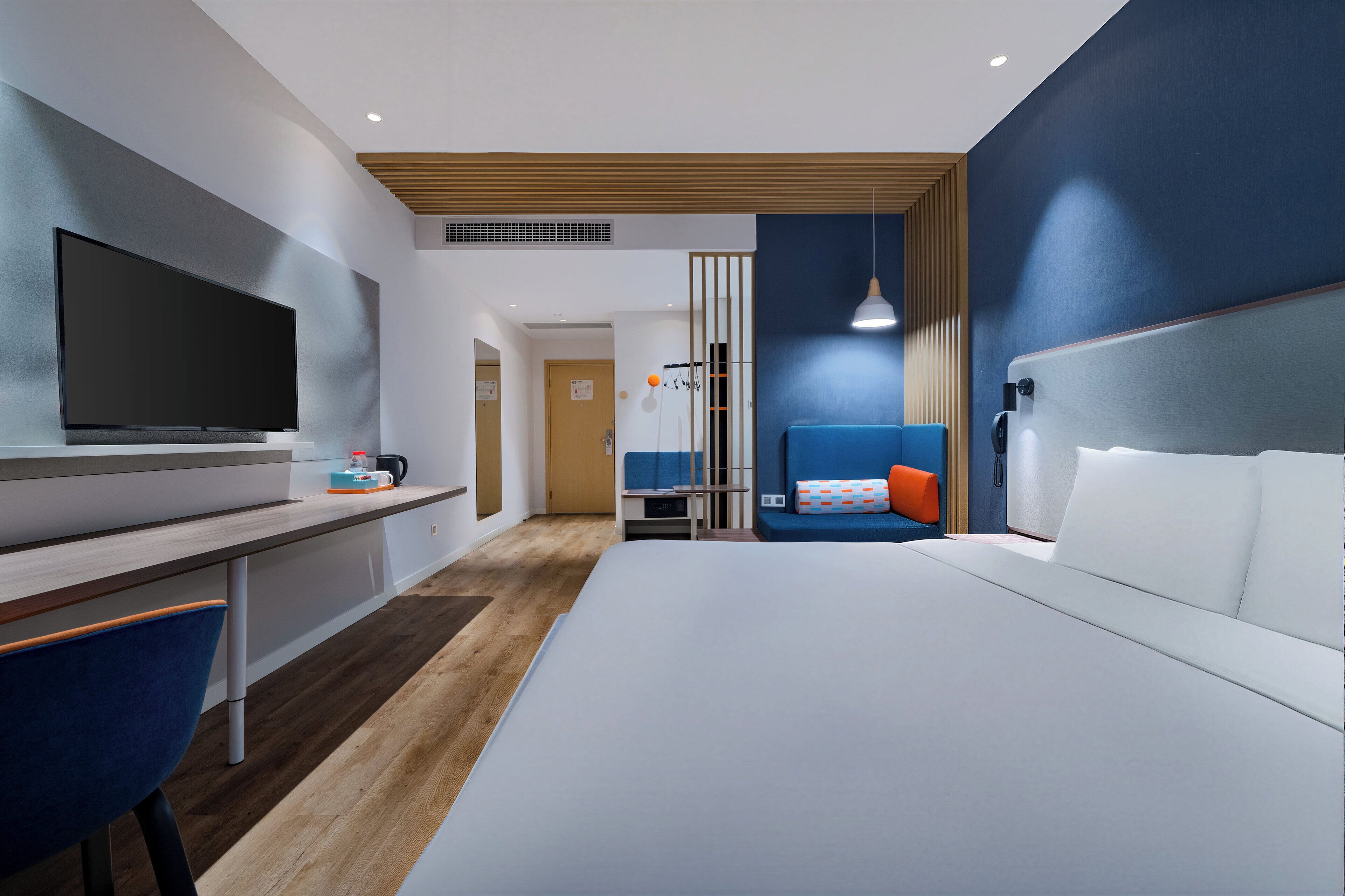 A modern hotel room with a large bed, a desk with a flat-screen TV, a blue chair, and a blue accent wall.