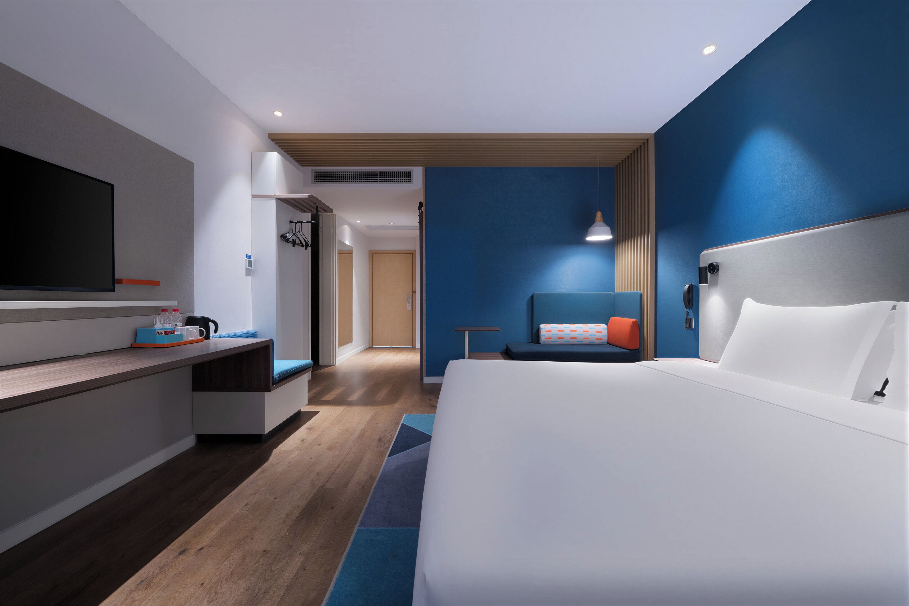 A modern hotel room with a large bed, a desk with a flat-screen TV, and a seating area with a blue chair and a small table.