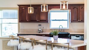 Una cocina moderna con armarios de madera, una isla central con un taburete blanco y mullido, luces colgantes y una planta en maceta.