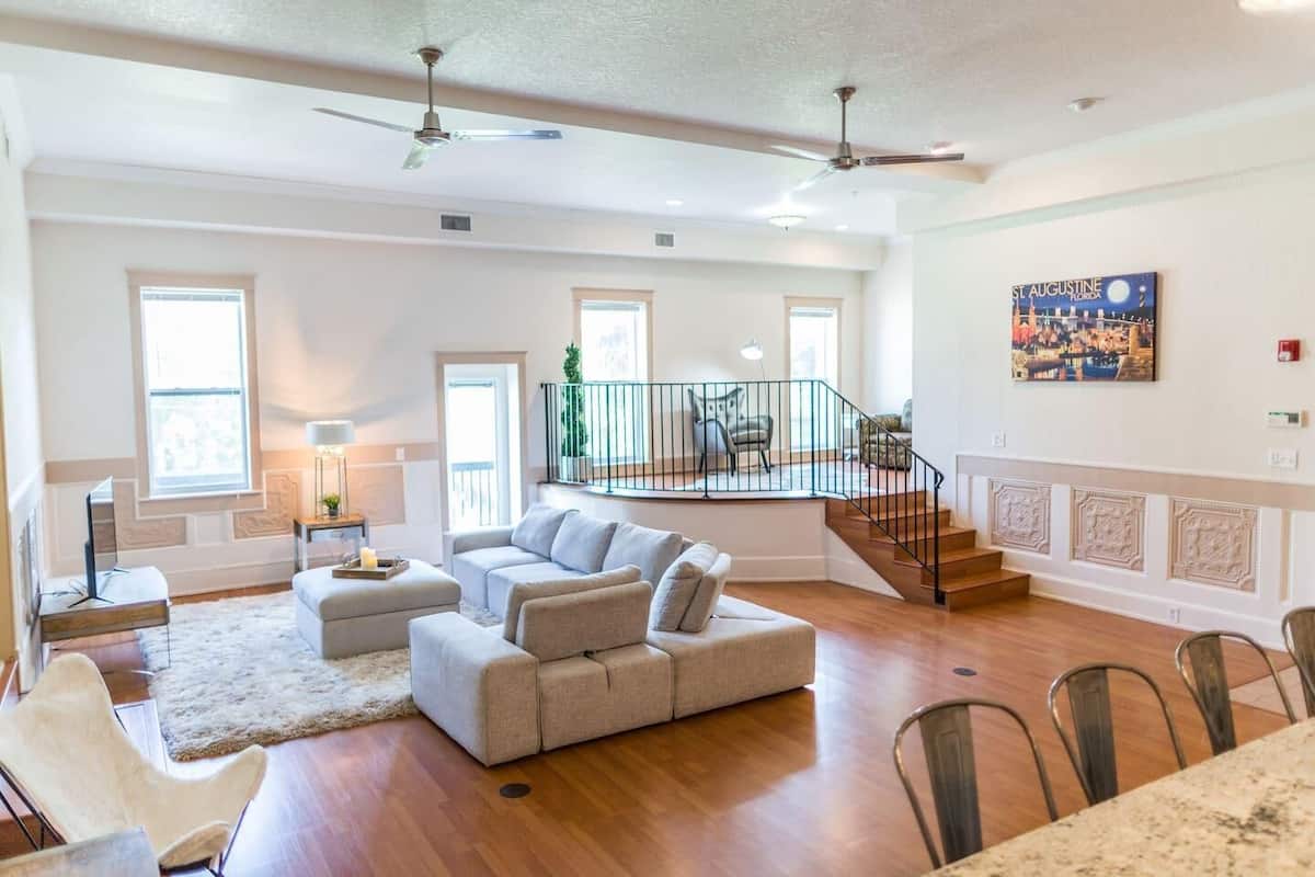 A spacious living room with a large sectional sofa, a dining area with a table and chairs, and a staircase leading to an upper level.