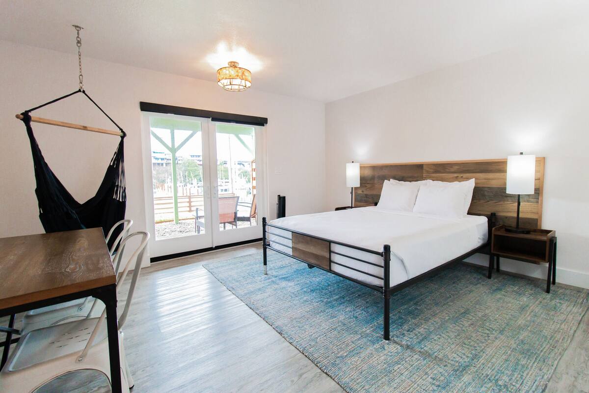 A hotel room with a bed, bedside tables, a hanging chair, a dining table, and a view of the outside.