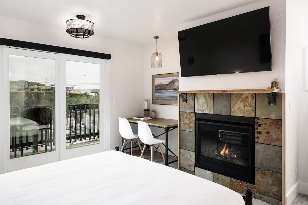 A modern bedroom with a large bed, a fireplace, a TV mounted on the wall, and a balcony with a view of the city.