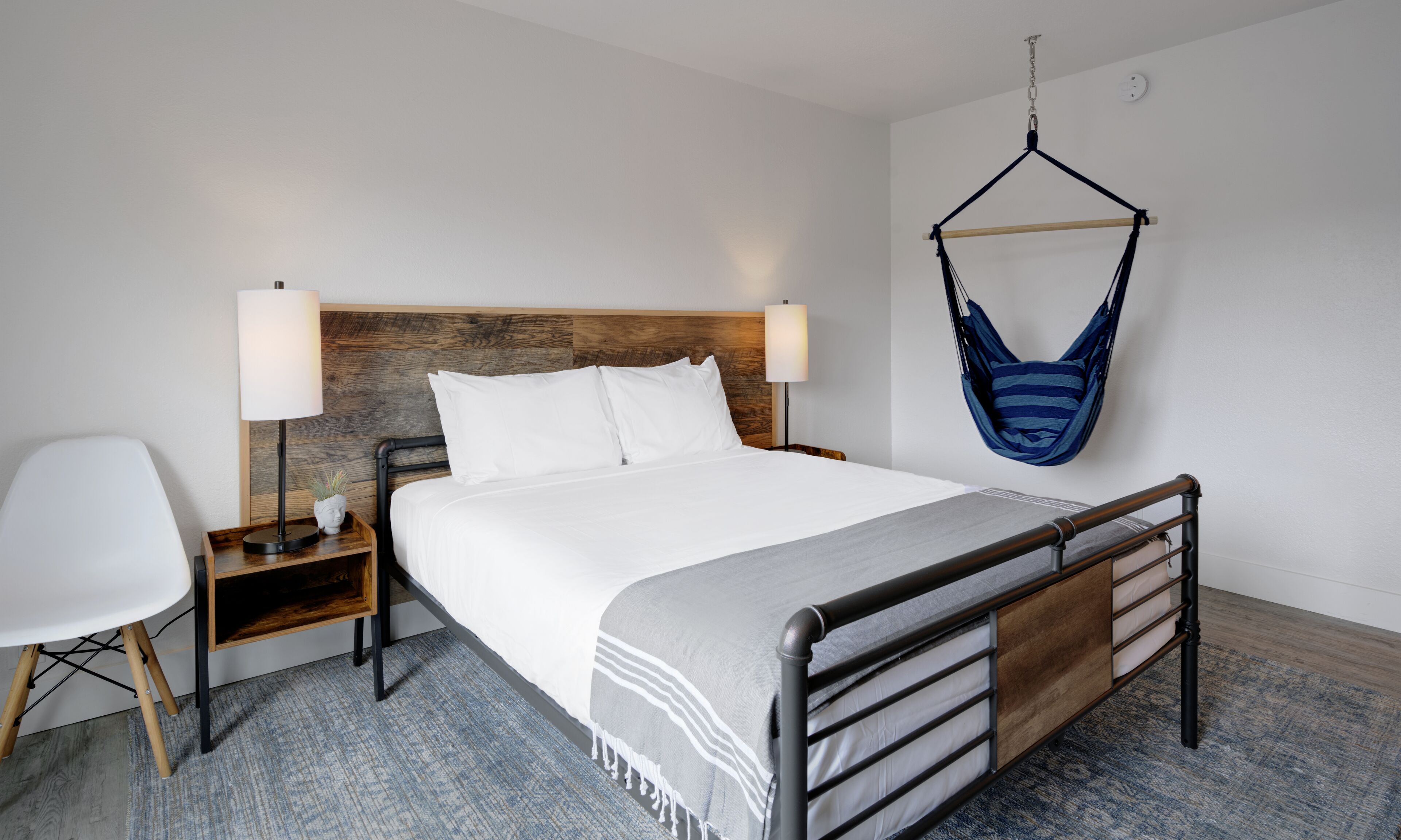 Une chambre d’hôtel moderne avec une tête de lit en bois, un lit recouvert de draps blancs, un hamac suspendu, une table de chevet avec une lampe et une chaise.