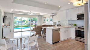 Cocina moderna con gabinetes blancos, lavavajillas integrado y horno de acero inoxidable. Hay un comedor con mesa y sillas blancas, y una vista a la piscina a través de amplias ventanas.