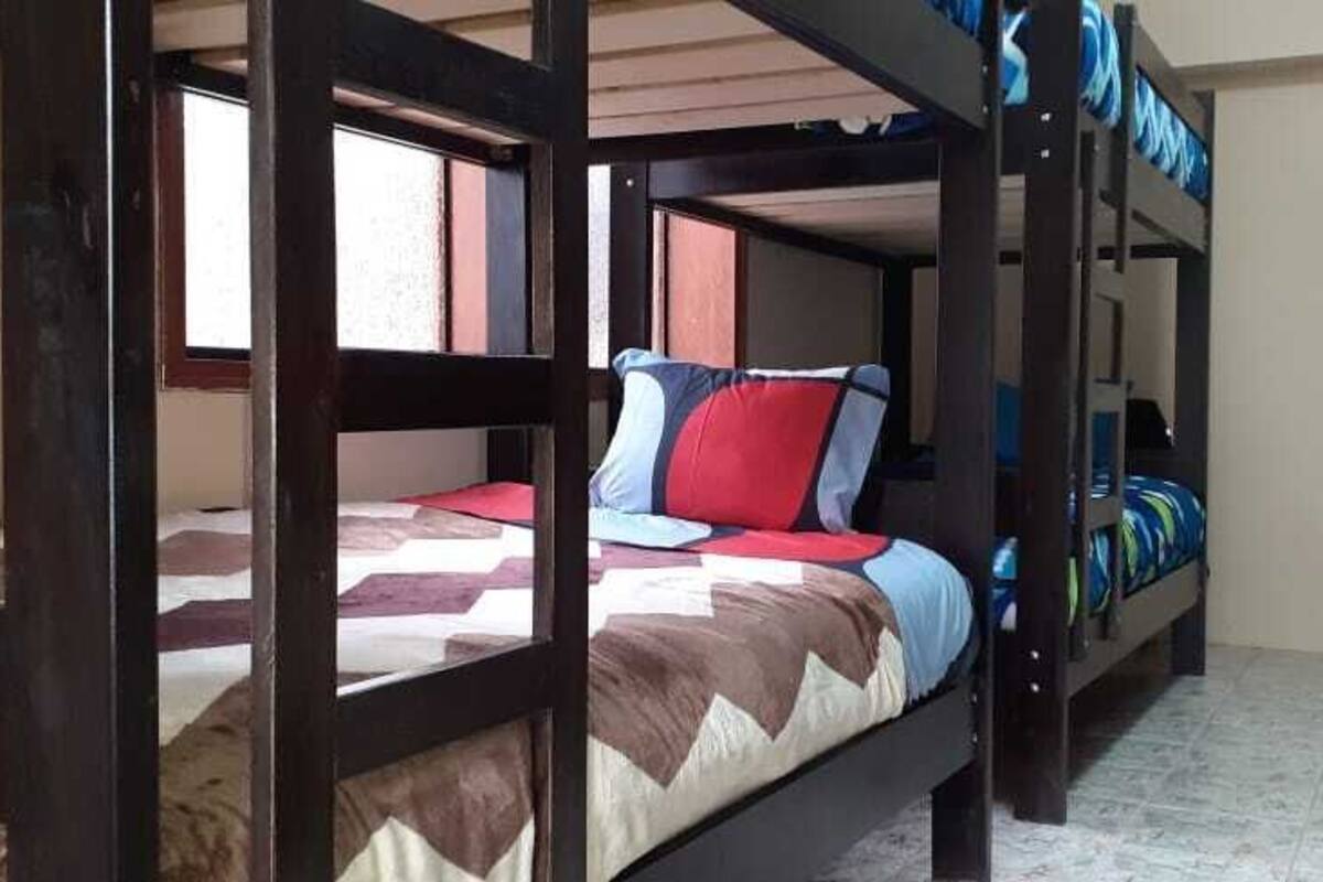 Ein Schlafraum mit Etagenbetten und Fenster, ein Bett mit gemustertem Bettbezug sowie eine Wand mit einer Tür.