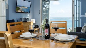 A dining table with two chairs, a wine bottle, and glasses of red wine. A TV is mounted on the wall, and a window offers a view of the sea. - A stunning apartment with breathtaking views, situated in a select development. (Carbis Bay)