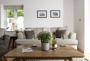 A living room with a sofa, two framed pictures on the wall, a coffee table with a magazine, a wine bottle, and glasses. - A stunning ground floor apartment with a large outdoor terrace and sea views. (Carbis Bay)